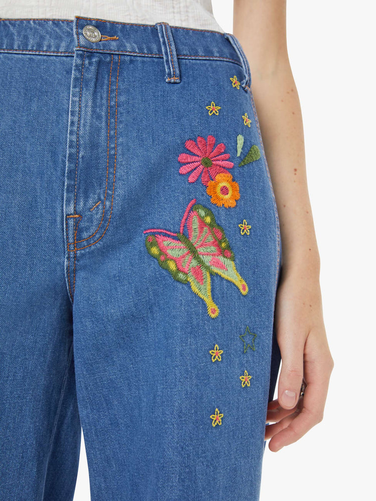 Close up view of a woman in a med blue with embroidery flowers  wide leg jeans with a high rise, zip fly, long inseam and a clean hem.