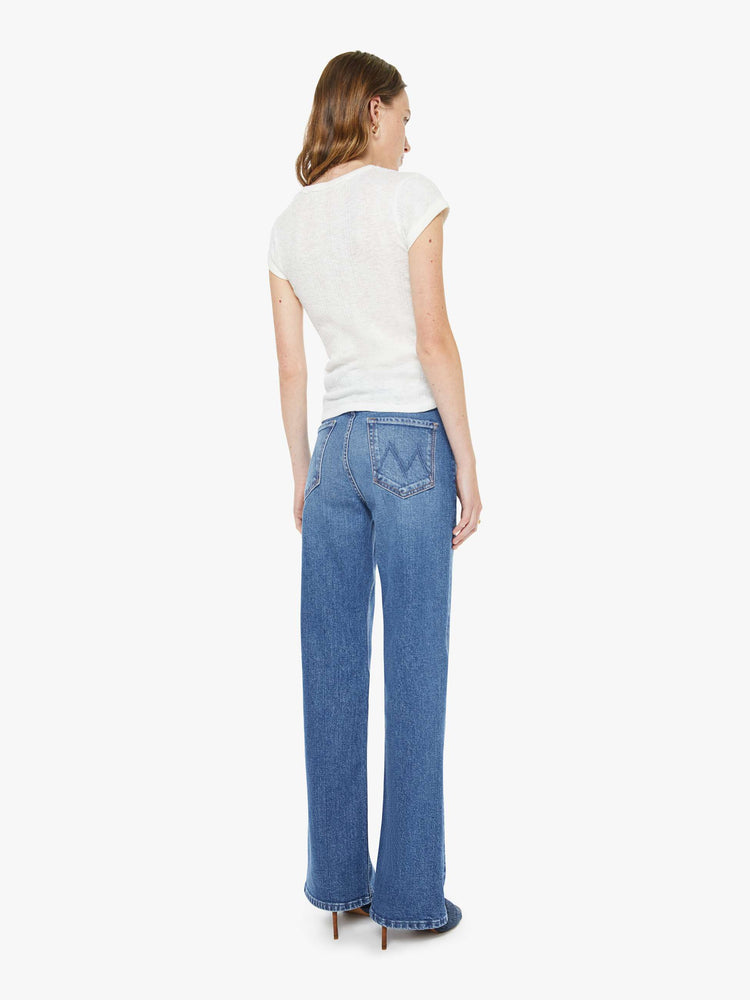Back view of a woman in a med blue high-rise bootcut with a zip fly, looser leg and a long 32-inch inseam with a clean hem.