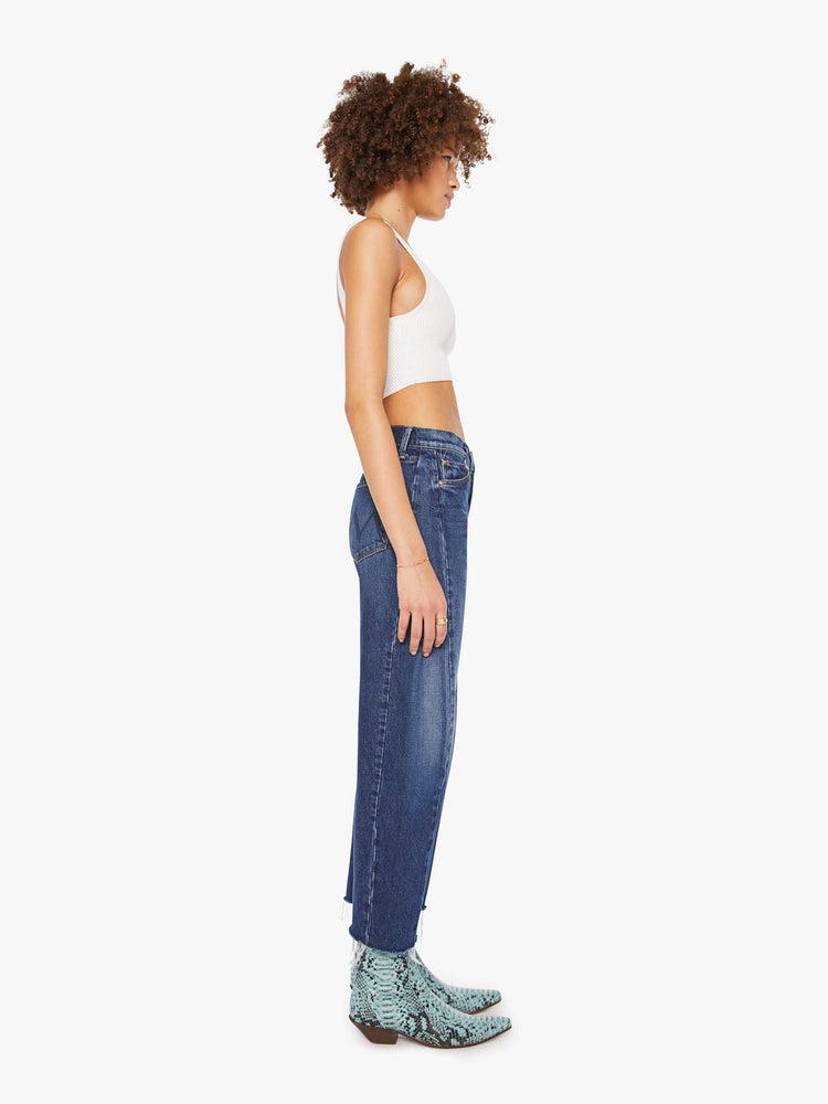 Side view of a woman in a dark blue mid-rise jeans designed to sit lower on the hips with a full barrel leg, zip fly, 28.25-inch inseam and a frayed hem.