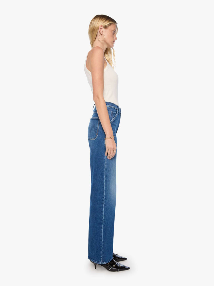 Side view of a woman in med blue super high-rise jeans with a loose fit, wide leg, patch pockets, zip fly and 34-inch inseam with a clean hem.