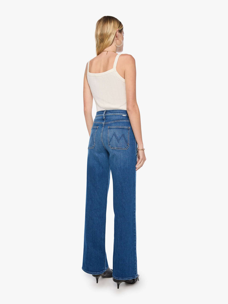 Back view of a woman in med blue super high-rise jeans with a loose fit, wide leg, patch pockets, zip fly and 34-inch inseam with a clean hem.