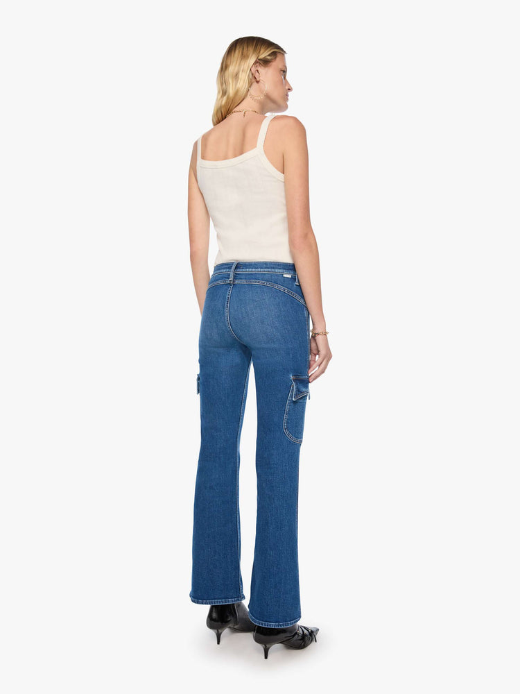 Back view of a woman in a med blue high-rise flare has a zip fly, patch pockets on the thighs, a curved seam across the back, 31-inch inseam and a clean hem. 