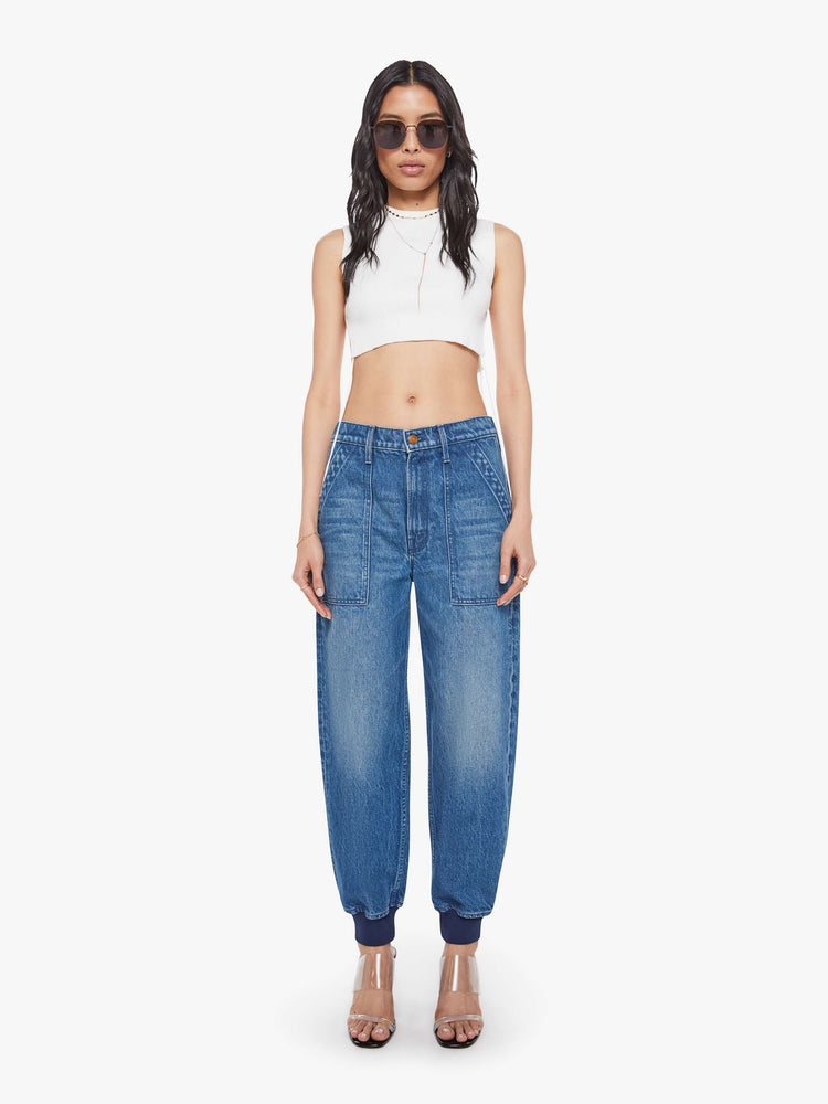 Front view of a woman in a mid blue  super high-waisted pants have a zip fly, slouchy fit designed to sit lower on the hips, oversized patch pockets and an ankle-length inseam with soft ribbed hems. 