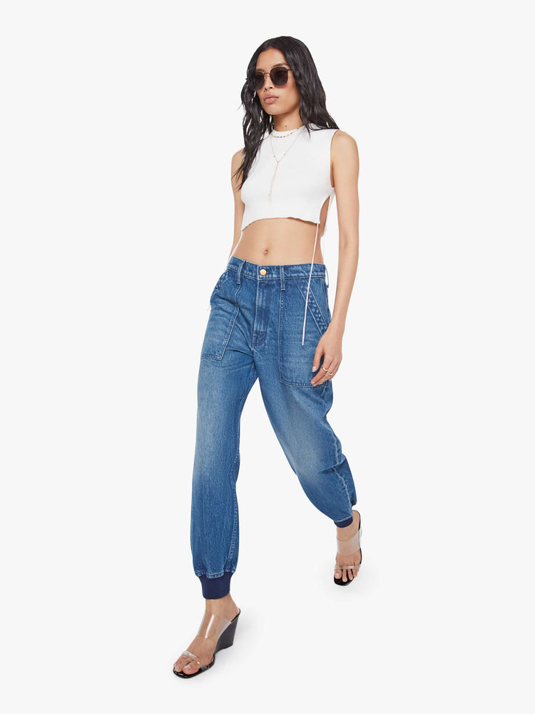 Walking view of a woman in a mid blue  super high-waisted pants have a zip fly, slouchy fit designed to sit lower on the hips, oversized patch pockets and an ankle-length inseam with soft ribbed hems. 