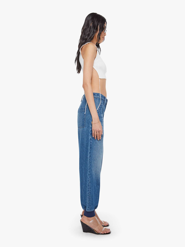 Side view of a woman in a mid blue  super high-waisted pants have a zip fly, slouchy fit designed to sit lower on the hips, oversized patch pockets and an ankle-length inseam with soft ribbed hems. 