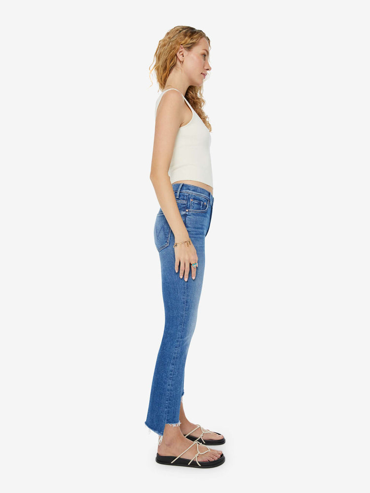 Side view of a woman in a high-rise mid-blue frayed step-hem bootcut jean with whiskering and fading at the knees. Styled with a white tank top and sandals. 