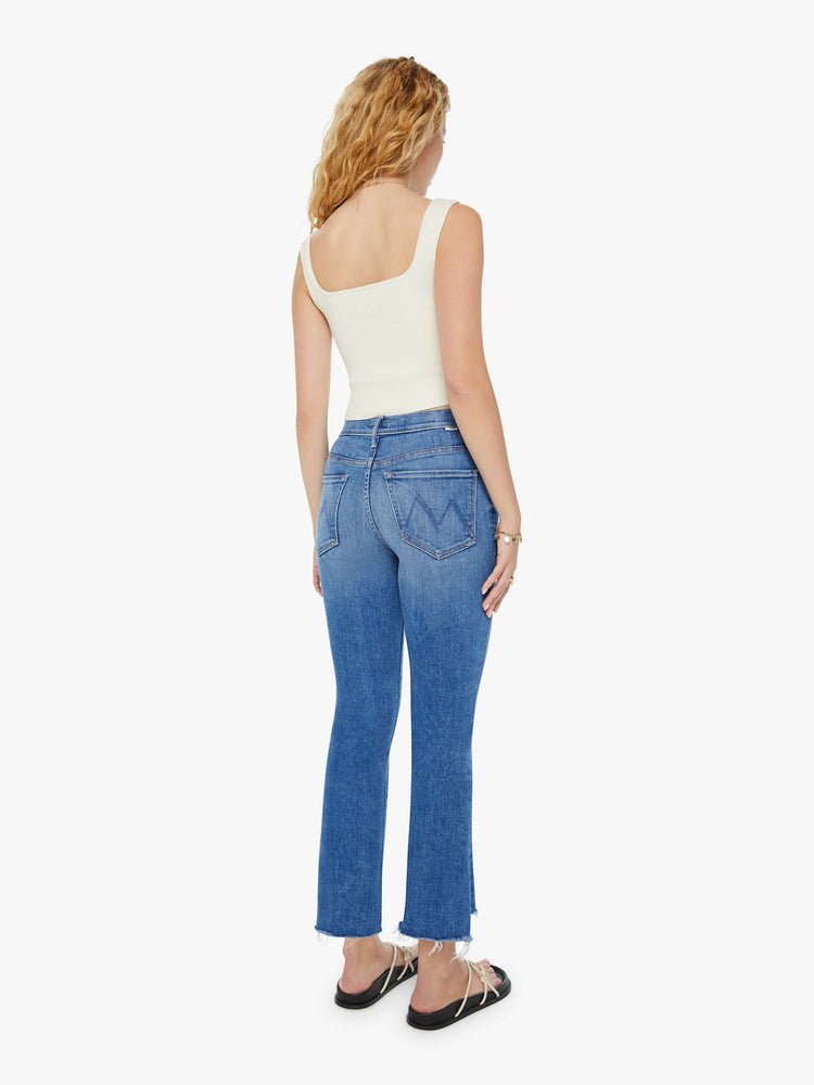 Back view of a woman in a high-rise mid-blue frayed step-hem bootcut jean with whiskering and fading at the knees. Styled with a white tank top and sandals. 