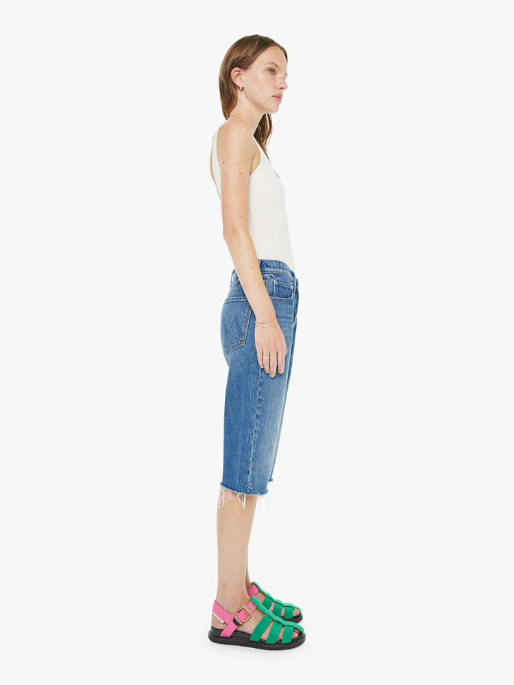Side view of a woman in a classic blue high-rise straight leg denim capri with a zip fly, 16-inch inseam and a frayed hem.