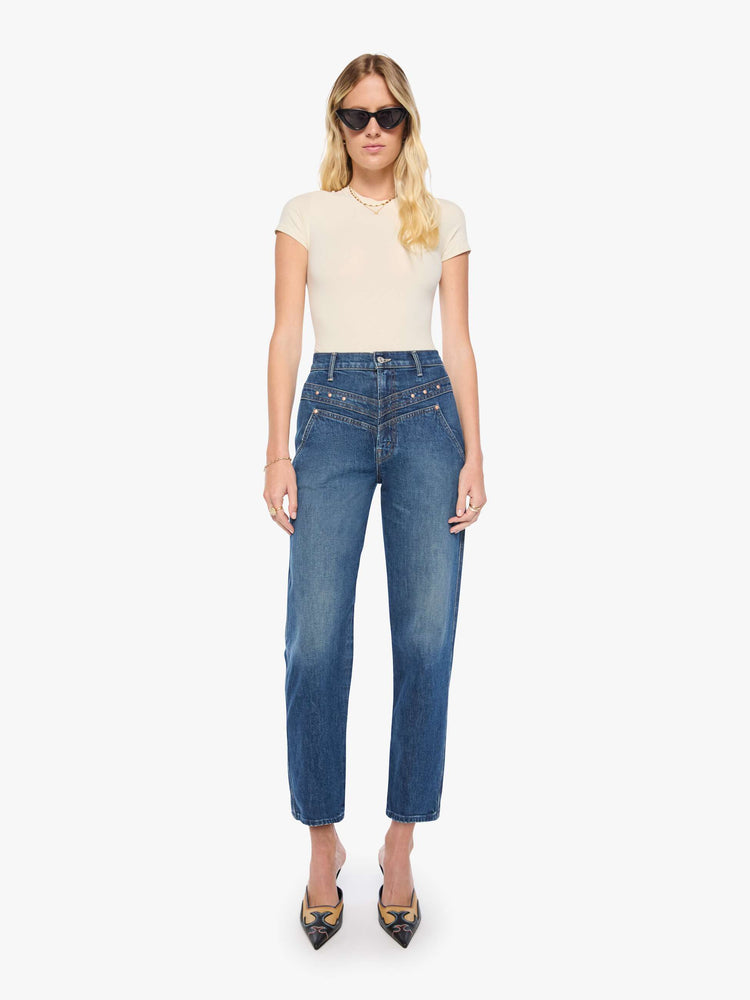 Front view of a woman in mid-blue with fading at the knees and rivet details on the front high-rise straight-leg jean with angled, Western-inspired seams at the waist, a zip fly, 29-inch inseam and a clean hem.