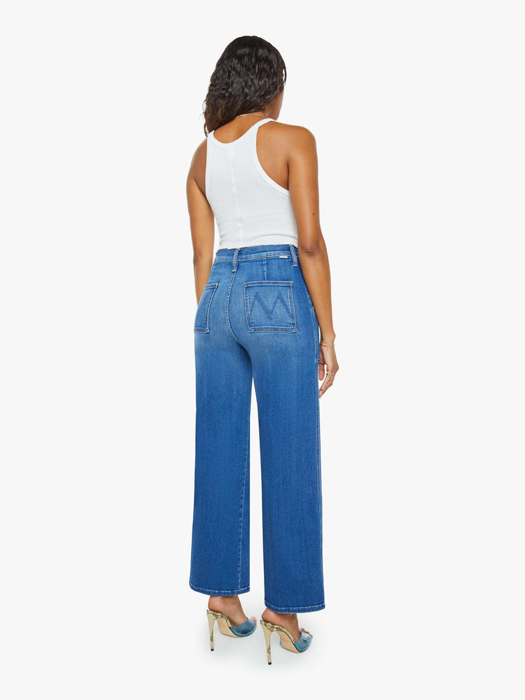 Back view of a woman in a mid blue super high-rise jeans with a loose fit, wide leg, zipper-fly, patch pockets and 29-inch inseam with a clean hem.