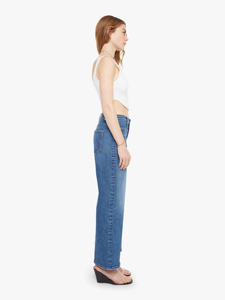 Side view of a woman in a med blue mid-rise jeans with a loose fit, zip fly, wide leg, patch pockets and ankle-length inseam with a clean hem.