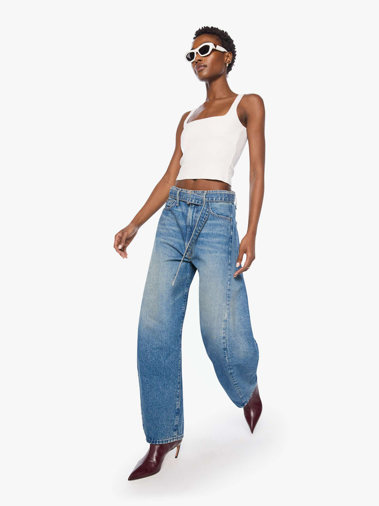 Walking view of a woman in a mid blue wide leg super high-waisted jeans are designed with a belted waistband, zip fly, an inset panel on the outer seam, slightly tapered leg and a clean hem.