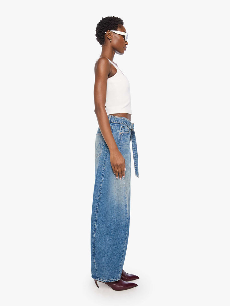Side view of a woman in a mid blue wide leg super high-waisted jeans are designed with a belted waistband, zip fly, an inset panel on the outer seam, slightly tapered leg and a clean hem.