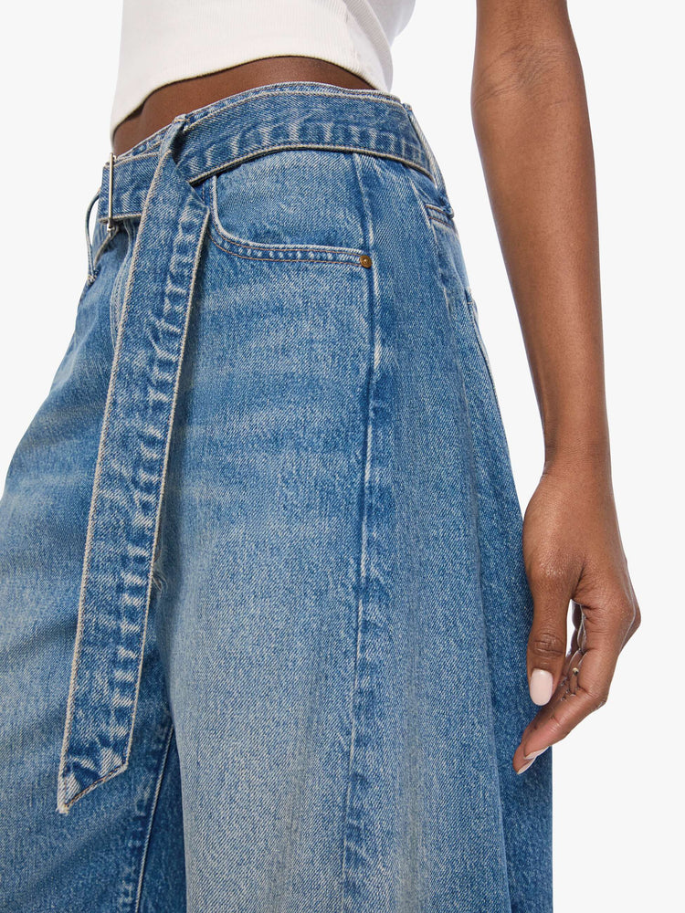 Close up view of a woman in a mid blue wide leg super high-waisted jeans are designed with a belted waistband, zip fly, an inset panel on the outer seam, slightly tapered leg and a clean hem.