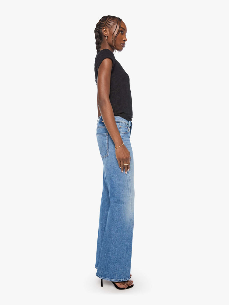 Side view of a woman in a mid blue mid-rise bootcut with a zip fly, looser leg, twisted outer seam and a long 31-inch inseam with a clean hem.