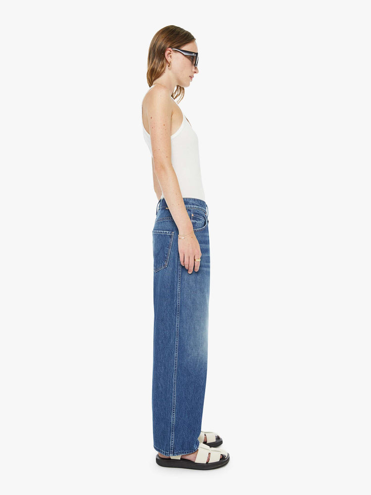 Side view of a woman in a mid blue jeans designed to sit lower on the hips with an ankle-length inseam, button fly, clean hem and slightly tapered leg. 