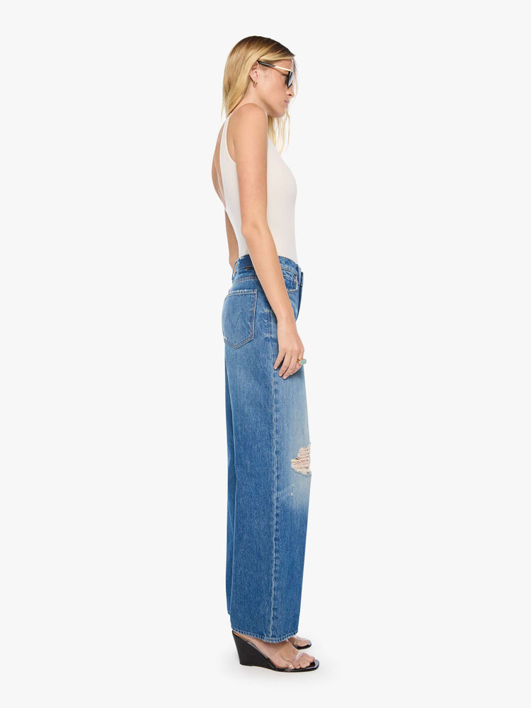 Side view of a woman in a med blue with tears in the knees high-waisted wide leg jeans with a button fly, 32-inch inseam and loose, low-slung fit. 