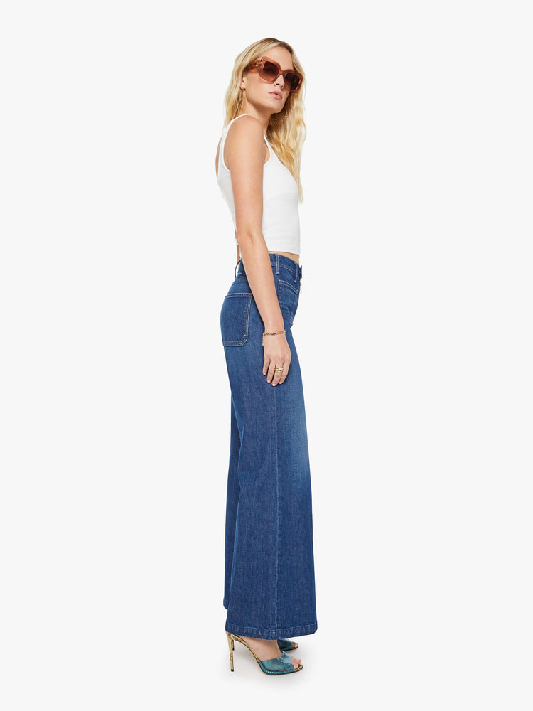 Side view of a woman in a dark blue wide leg jeans with a high rise, exposed zip fly, tabbed waistband, patch pockets and a long 34-inch inseam with a clean hem.