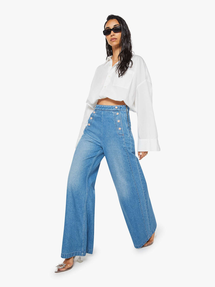 Walking view of a woman in a med blue super high-waisted jeans have a loose, wide leg, sailor-inspired buttons on the front, a left side zipper and a long 32-inch inseam with a clean hem.