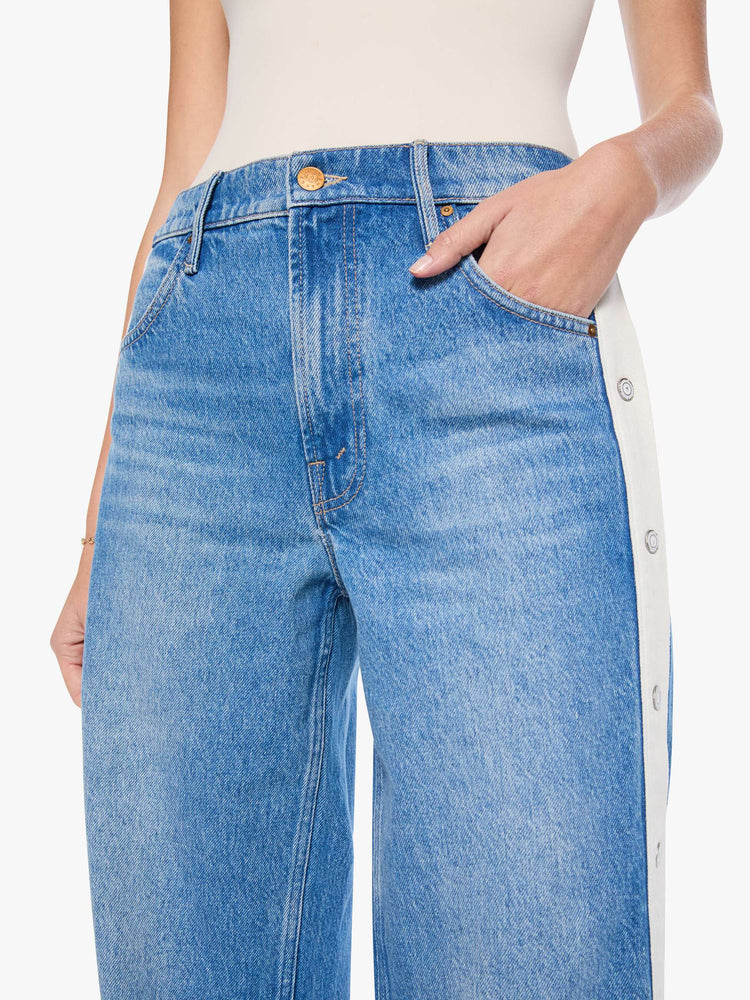 Close up view of mid blue high-waisted jeans have a loose, low-slung fit, zip fly, a white stripe down the outer seam with adjustable snaps, a long 31-inch inseam and a clean hem.
