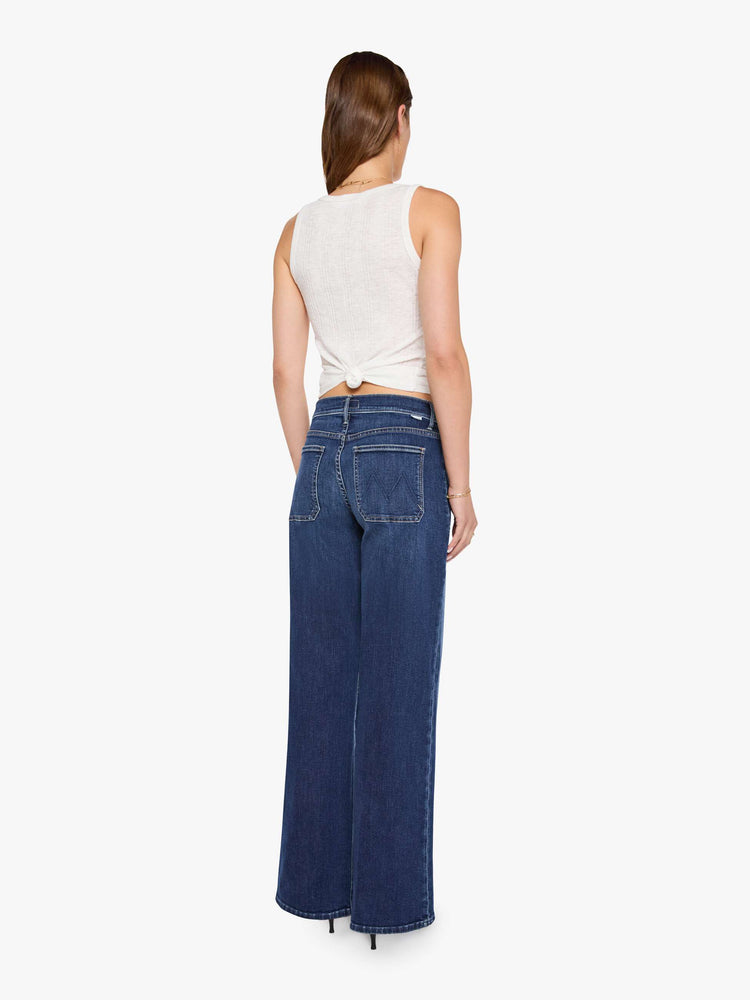 Back view of a woman in a dark blue mid-rise jeans with a loose fit, wide leg, patch pockets, zip fly and 32-inch inseam with a clean hem.
