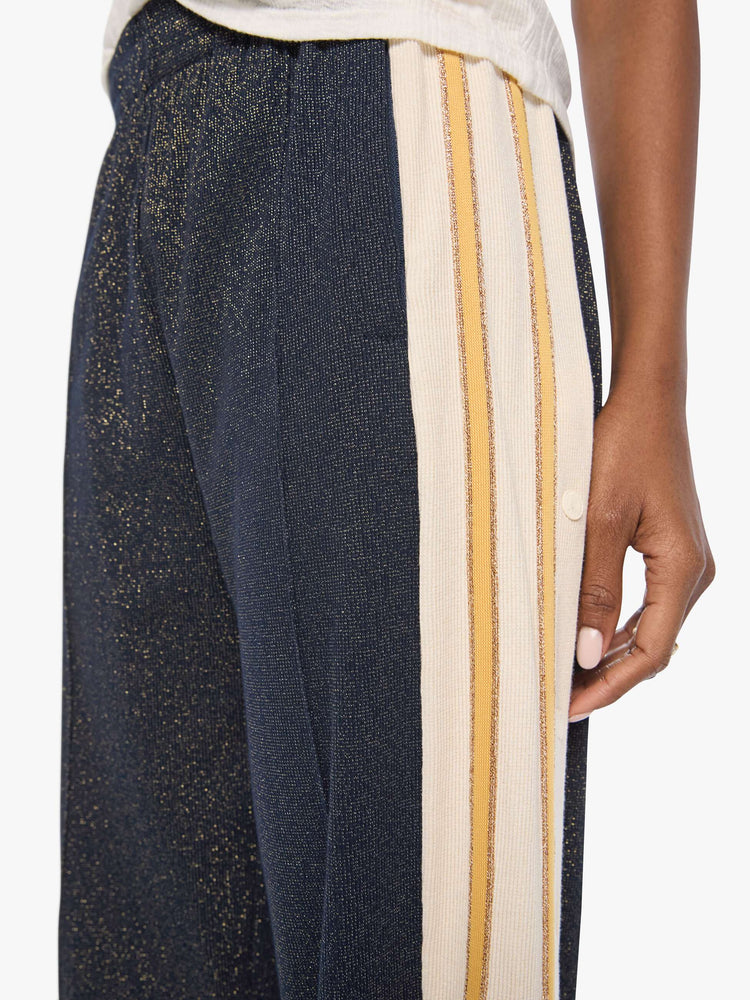 Close up  view of a woman in a metallic navy with cream and golden yellow stripes down the legs wide-leg track pants with an elastic waistband, loose leg and a contrast side stripe with buttons down the outer seams. 