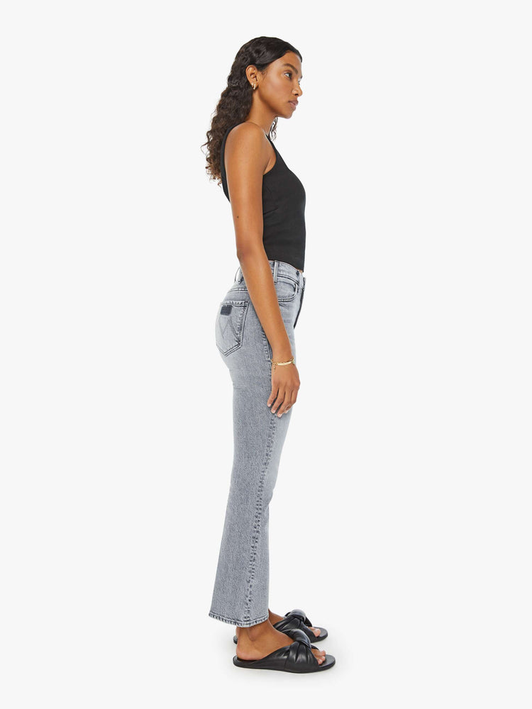 Side view of a woman in light-grey high-rise flare that has an ankle-length inseam and a clean hem with whiskering and fading. Paired with a black tank top and sandals.