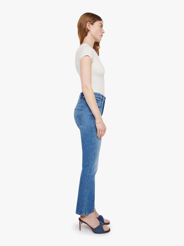 Side view of a woman in a mid blue high-rise bootcut has an ankle-length inseam, zip fly and a clean hem.