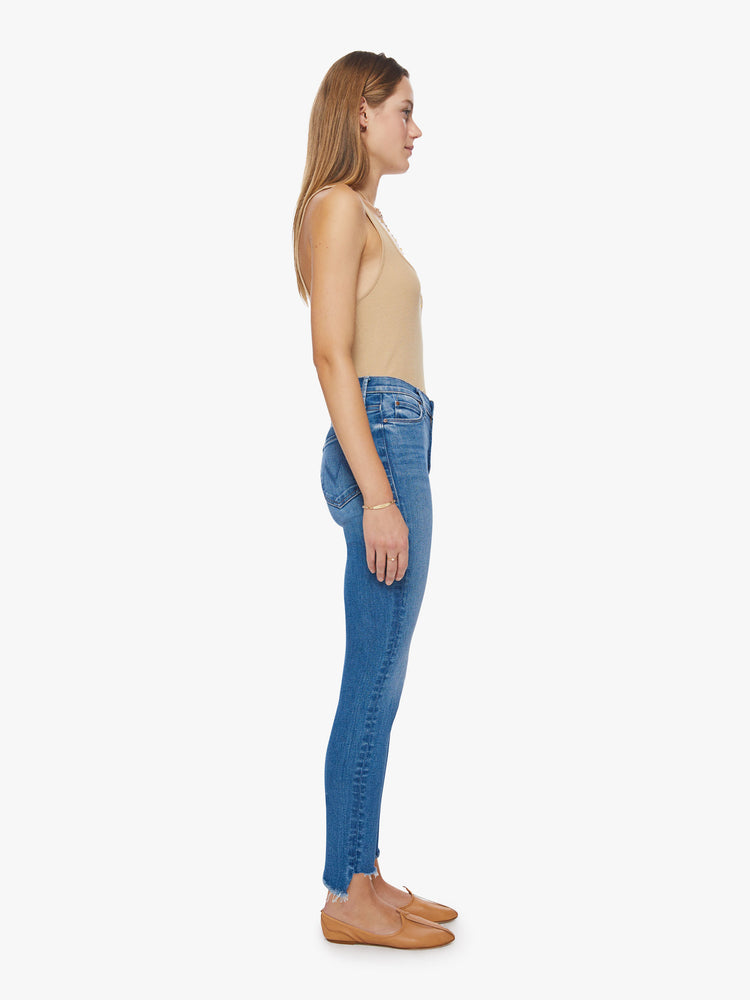 Side view of a woman high-rise skinny jean has a zip fly, ankle-length inseam and a frayed step-hem in a mid-blue wash.