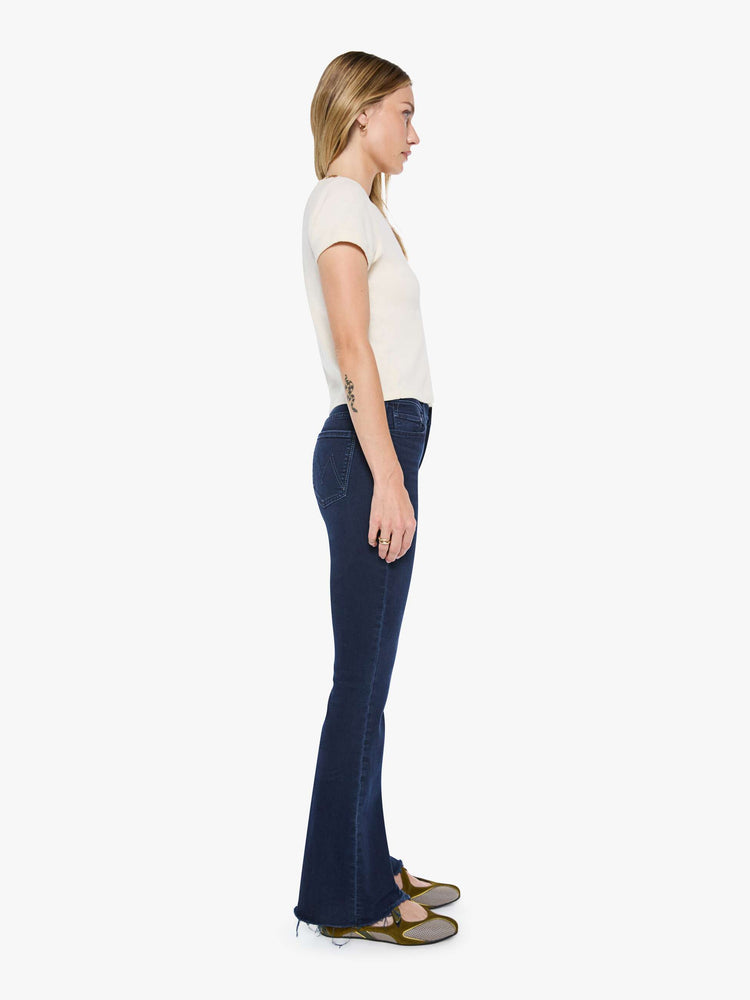 Side view of a woman in a dark blue wash mid-rise flare with a 31-inch inseam and frayed hem.
