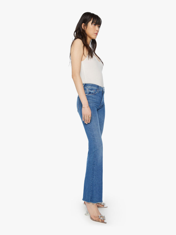 Front angle view of a woman wearing medium blue wash denim featuring a mid rise and an ankle length flare leg with a raw cut hem.