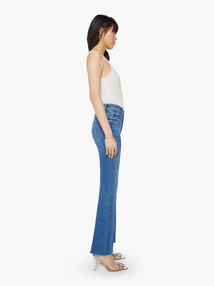 Side view of a woman wearing medium blue wash denim featuring a mid rise and an ankle length flare leg with a raw cut hem.