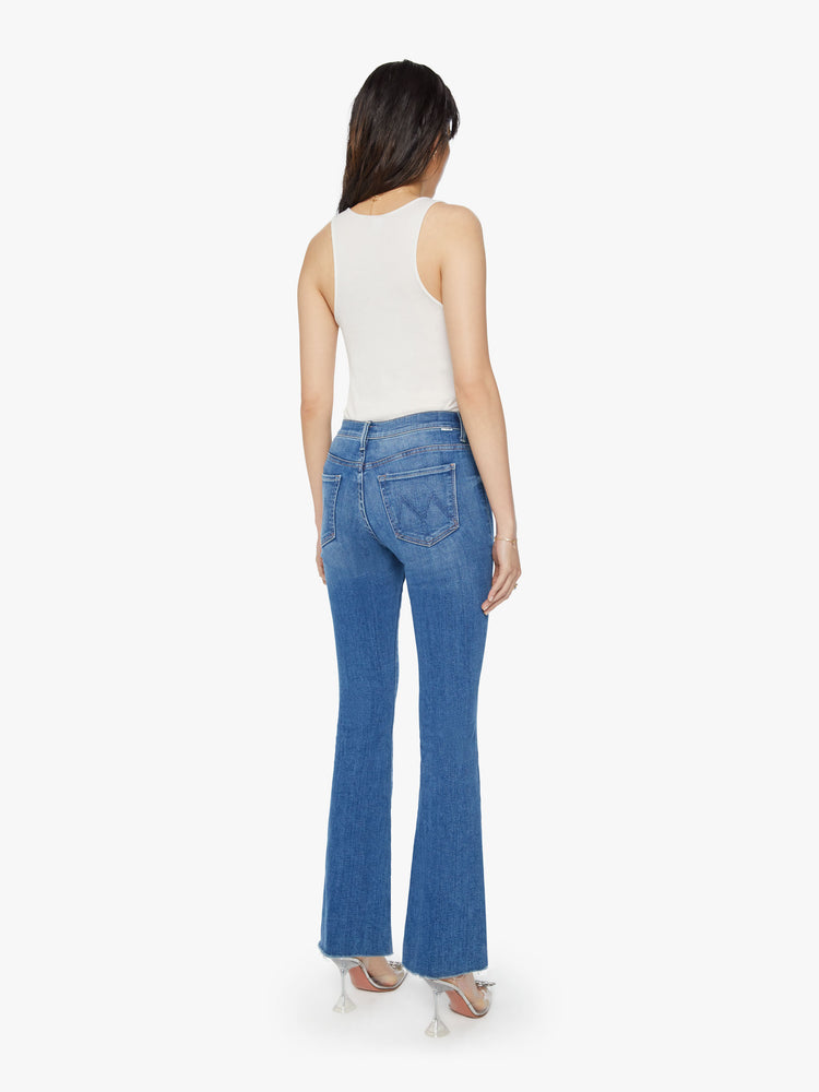 Back view of a woman wearing medium blue wash denim featuring a mid rise and an ankle length flare leg with a raw cut hem.