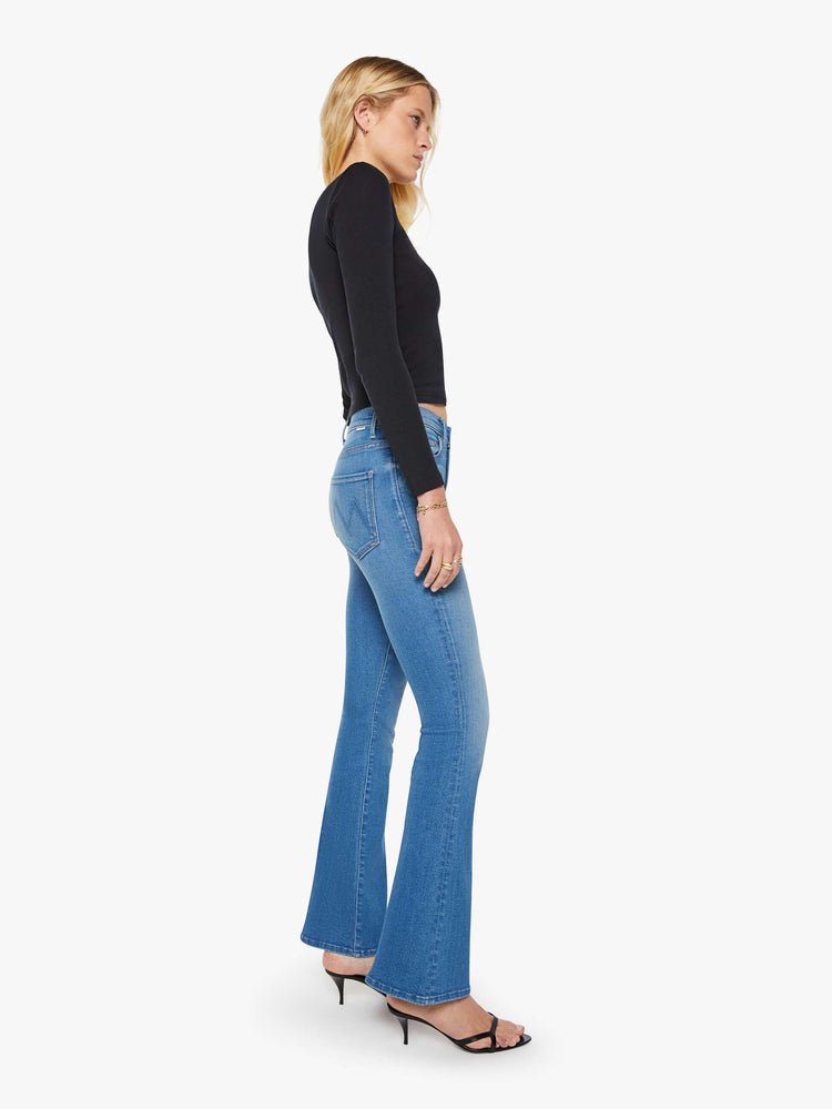 Side view of a woman in a med blue flare jean has a mid rise with a 31-inch inseam and a clean hem.
