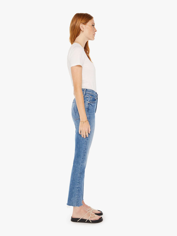 Side view of a medium blue wash jean featuring a high rise, a straight leg, and angle length fray hem.