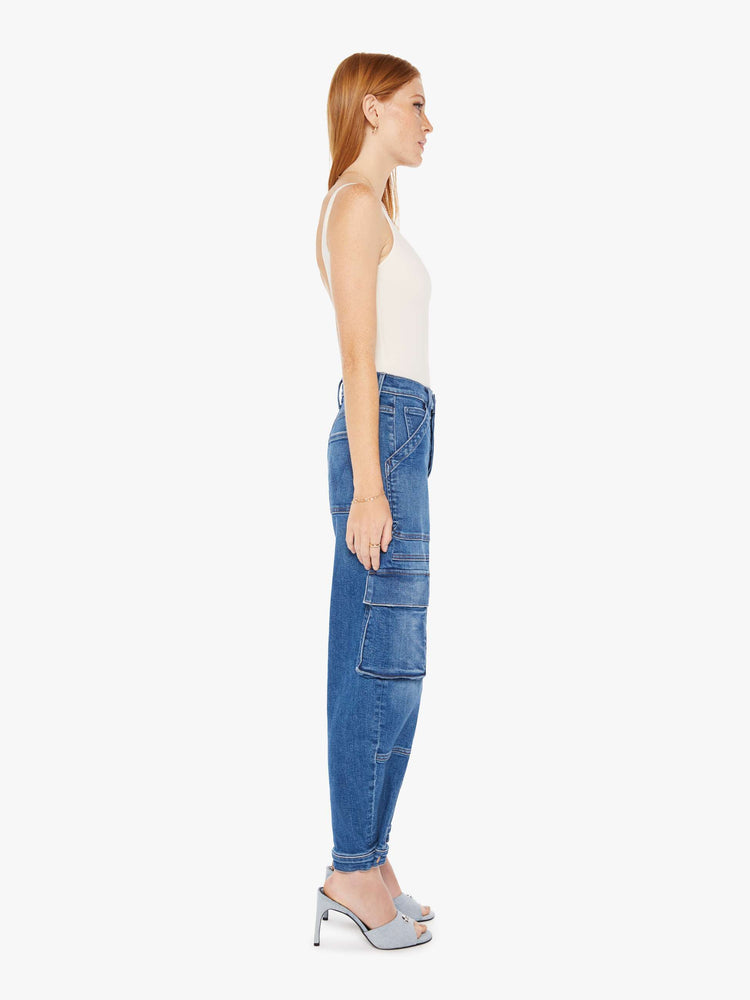 Side view of a womens medium blue wash cargo jean featuring a high rise and a tapered hem with snap buttons.