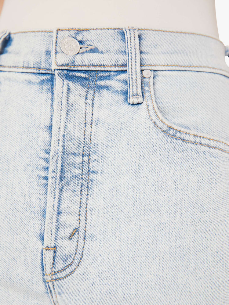 A close up swatch detail view of a light blue wash jean with darker blue hues around the seams, white hardware, and contrast beige stitching.