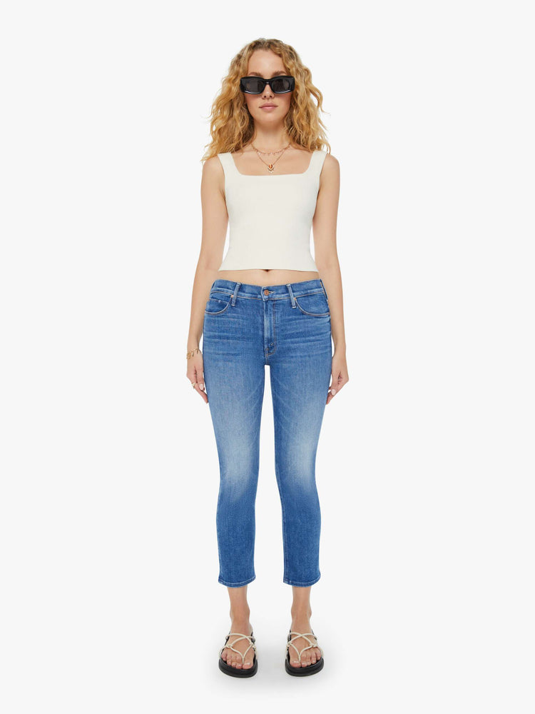 Front view of a woman in mid-blue straight leg jeans with a cropped hem and whiskering and fading at the knees. Styled with a white tank top. 