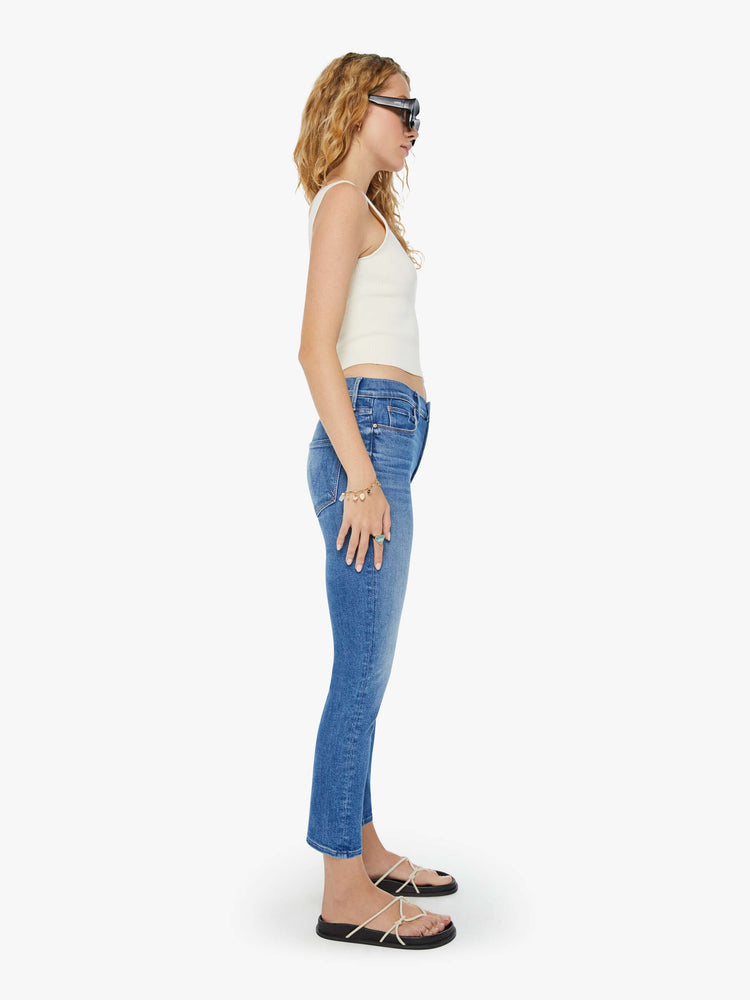 Side view of a woman in mid-blue straight leg jeans with a cropped hem and whiskering and fading at the knees. Styled with a white tank top. 