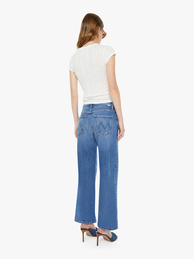 Back view of a woman in a mid blue jeans designed to sit lower on the hips with a zip fly, loose wide leg and a clean ankle-length inseam. 