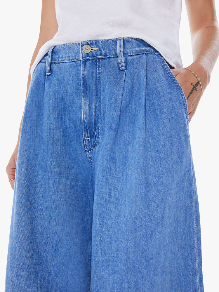 Close up view of a women high-waisted wide-leg pants with an ankle-length inseam and pleats at the waist in a mid-blue wash with faded details.
