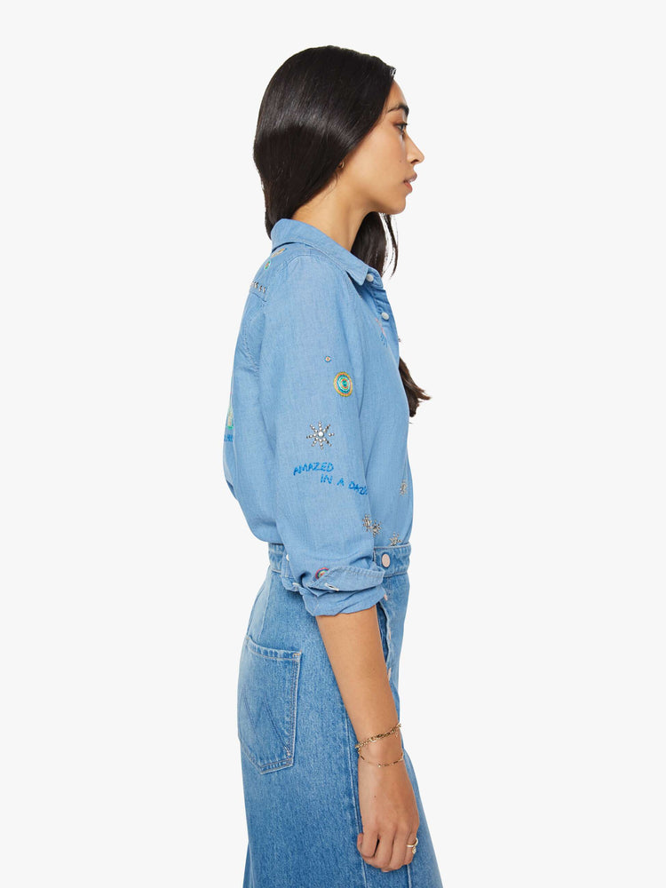 Side view of woman in a blue with colorful embroidery button-up blouse with patch pockets and a curved hem.