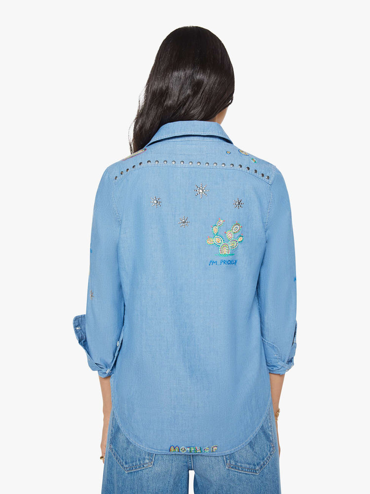 Back view of woman in a blue with colorful embroidery button-up blouse with patch pockets and a curved hem.