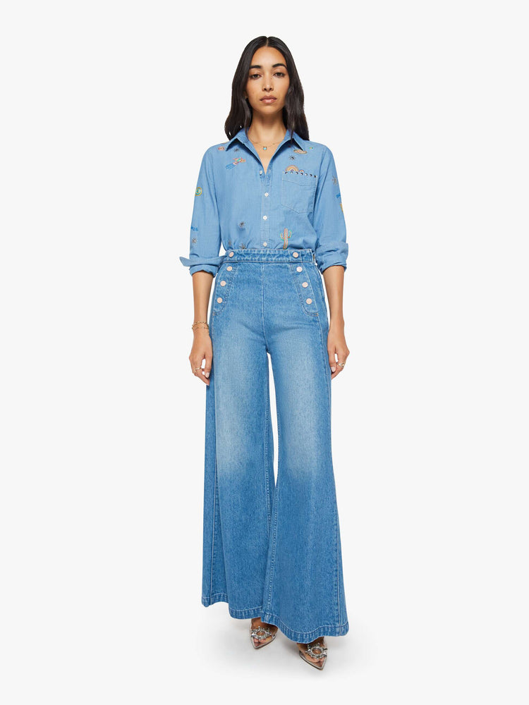 Full body view of woman in a blue with colorful embroidery button-up blouse with patch pockets and a curved hem.