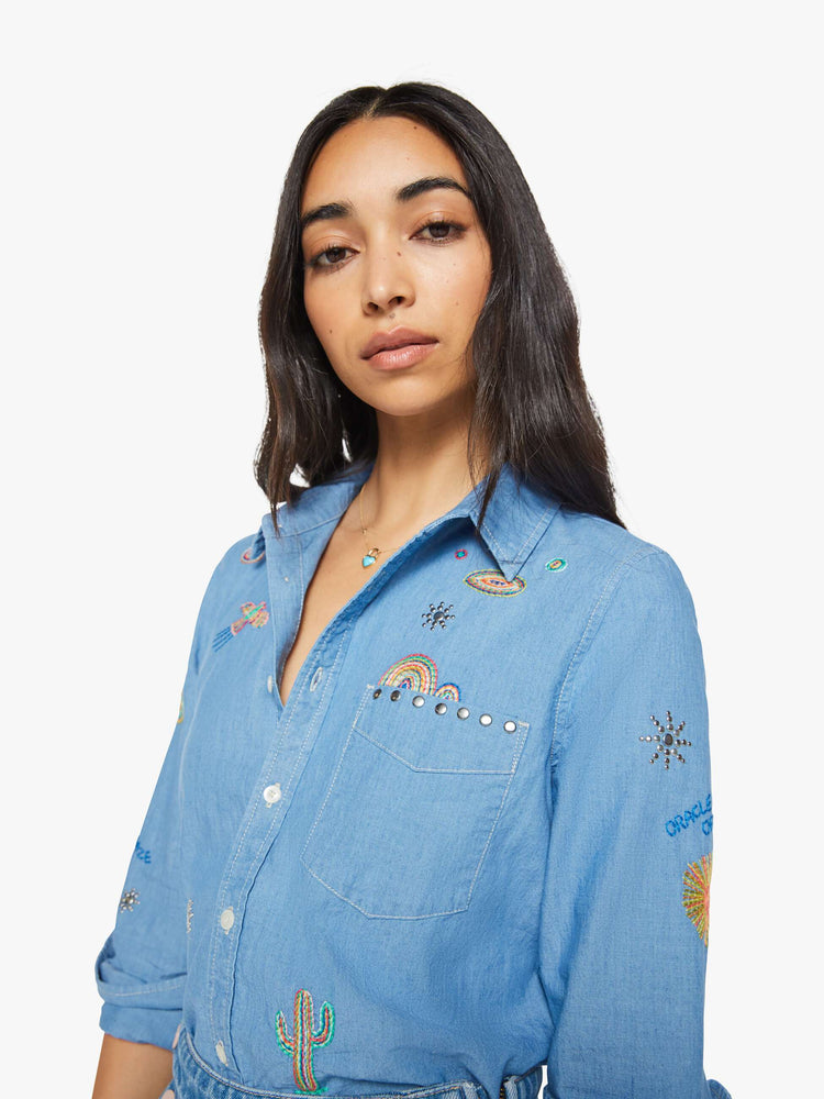 Close up view of woman in a blue with colorful embroidery button-up blouse with patch pockets and a curved hem.