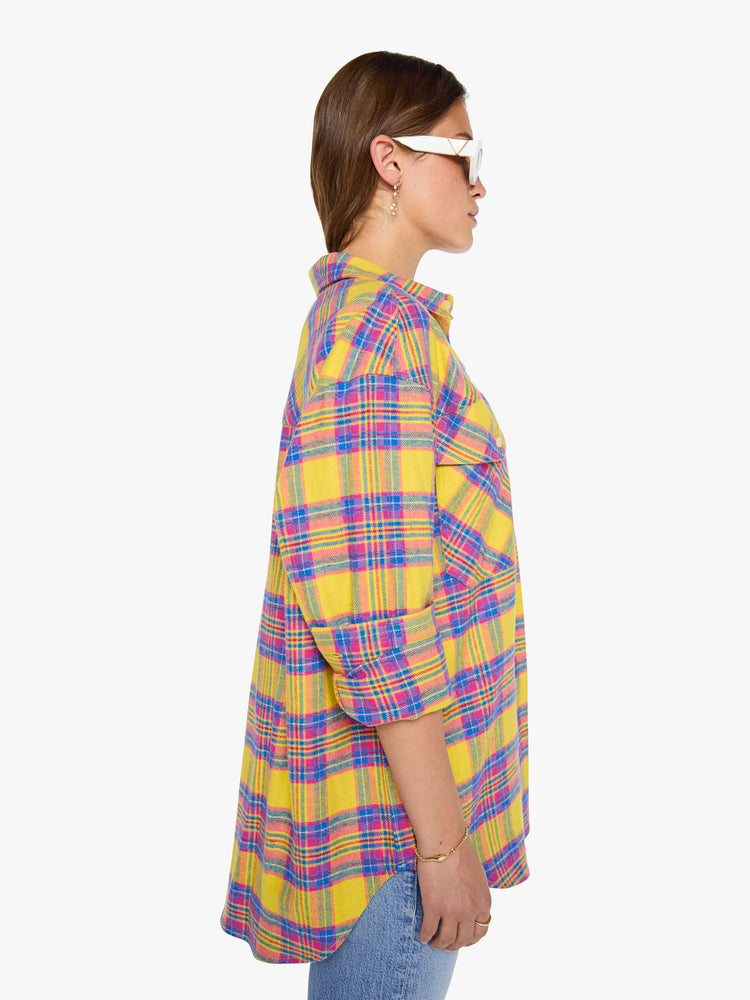 Side view of a woman in a yellow, pink and blue plaid print oversized button-up top with front patch pockets and a curved hem.