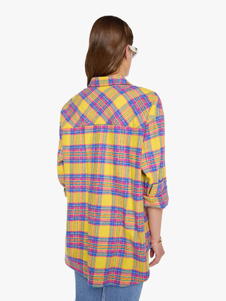Back view of a woman in a yellow, pink and blue plaid print oversized button-up top with front patch pockets and a curved hem.
