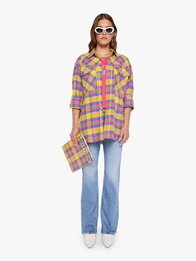 Full body view of a woman in a yellow, pink and blue plaid print oversized button-up top with front patch pockets and a curved hem.