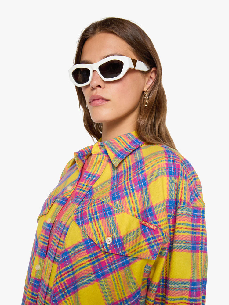 Close up view of a woman in a yellow, pink and blue plaid print oversized button-up top with front patch pockets and a curved hem.