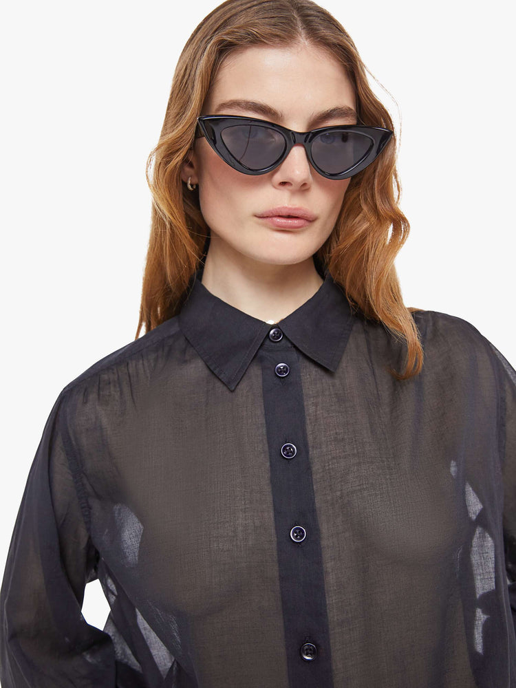Close up view of woman in a sheer black  collared button-up blouse with long balloon sleeves, a curved hem and an oversized fit. 
